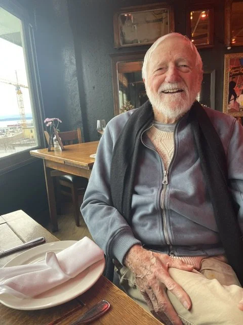 An elderly man with a beard smiling at a restaurant table, with a window and artwork in the background.