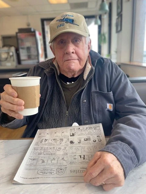 An elderly man wearing a beige cap and a dark jacket sitting at a table in a cafe, holding a coffee cup and looking at the camera.