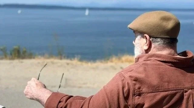 Older man with gray hair and beard wearing a tan hat and brown shirt, sitting outdoors near a body of water with sailboats in the background.
