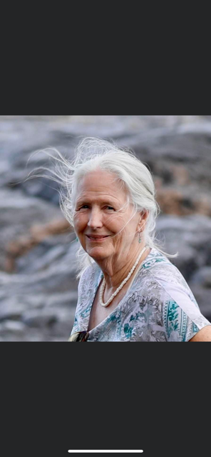 An elderly woman with white hair standing outdoors near a body of water, smiling at the camera, with a rocky background and wearing a floral top and pearl necklace.