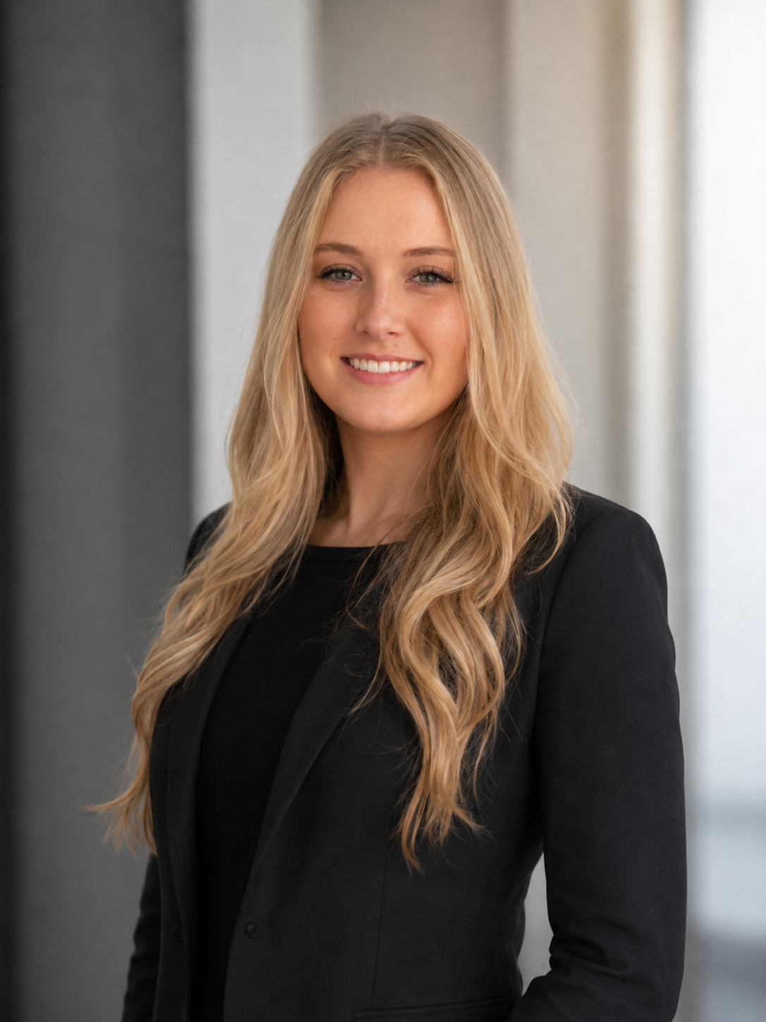 Professional portrait of a young woman with long blonde hair, smiling, wearing a black blazer, standing indoors with blurred background.