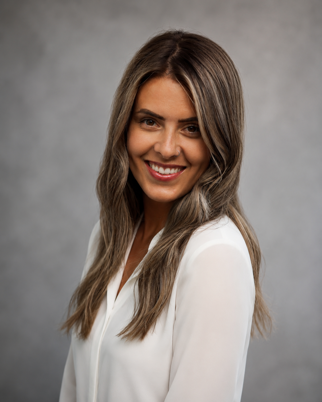 A woman with long layered hair smiling, wearing a white blouse, against a gray background.
