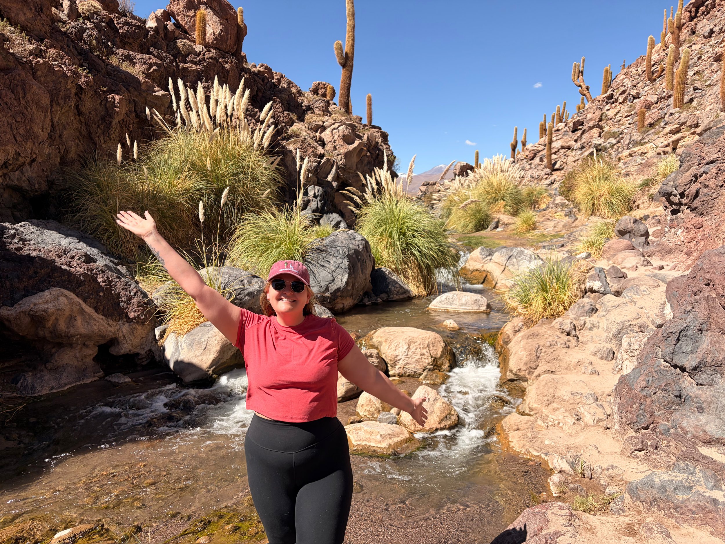 Piper, founder of Tern & Meridian, smiling with one arm raised beside a rushing stream lined with pampas grass and cacti in the Atacama Desert.