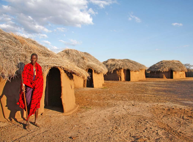 A man in traditional Maasai clothing standing outside mud and thatch huts in a village with a dirt ground and blue sky.