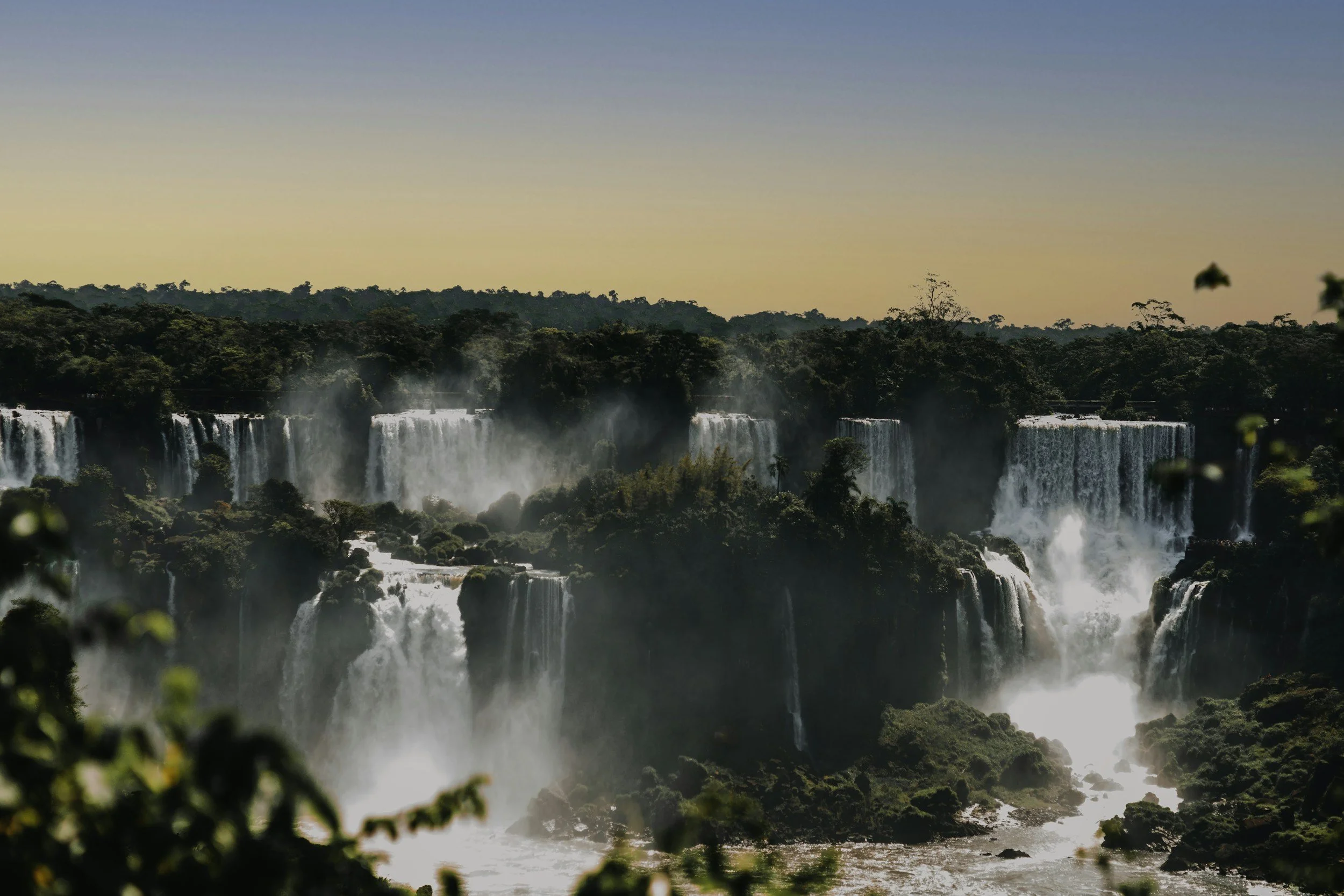 Waterfalls cascading over rocks with lush green forest in the background during sunset.