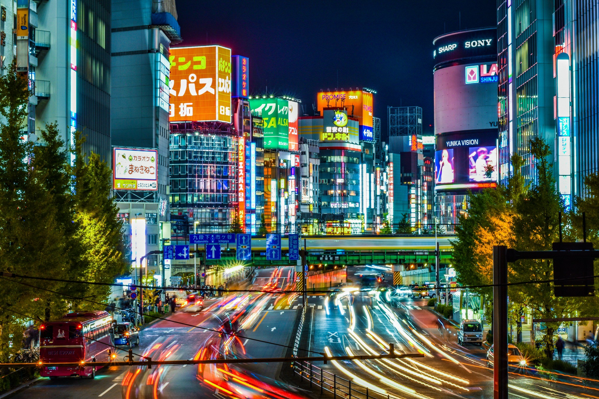 Nighttime city street filled with colorful lights and neon signs, with blurred moving vehicles creating light trails, tall buildings covered in illuminated advertisements, and trees lining the road.