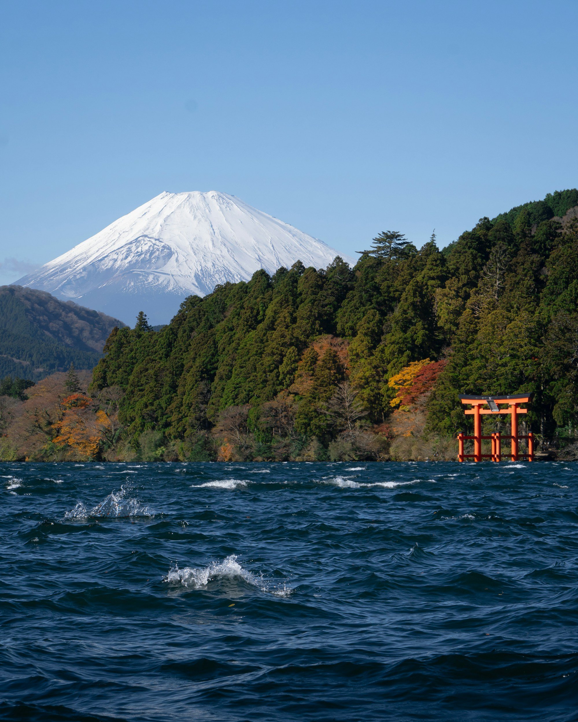 A view of Mount Fuji with snow-capped peak, green forested hills, and a torii gate in the water in the foreground.