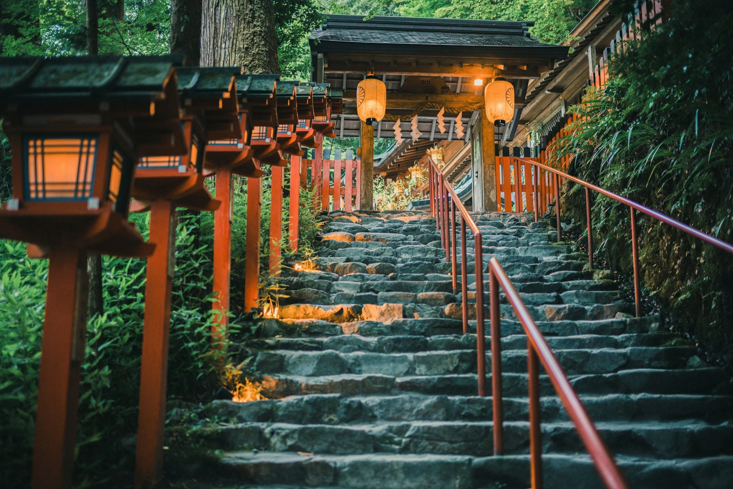 Stone steps leading up through a lush green forest with traditional lanterns hanging along the pathway in a Japanese shrine or temple setting.