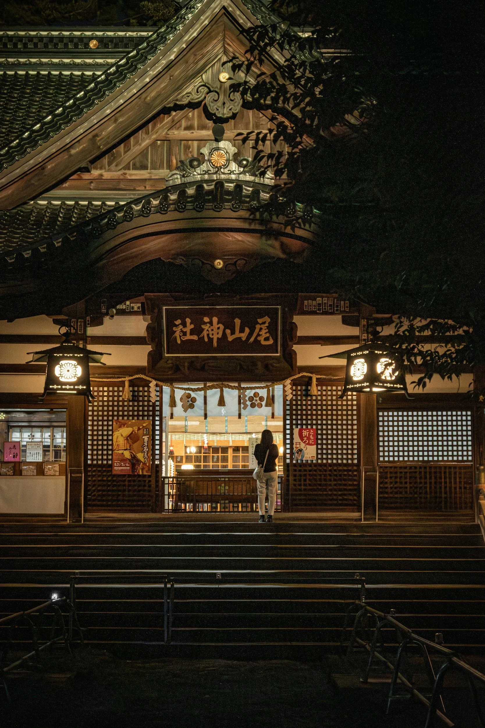 Night view of a traditional Japanese shrine entrance with wooden architecture, lanterns, and a person standing in front, illuminated softly in the dark.
