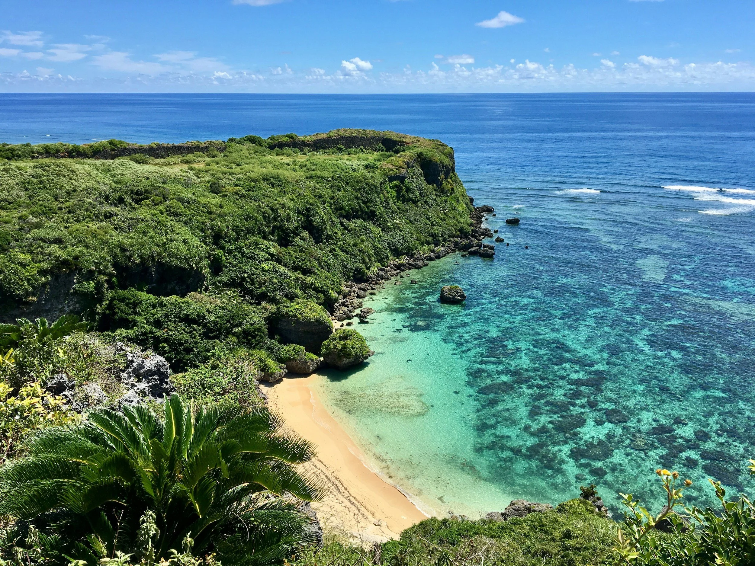 A lush green tropical cliff overlooking a sandy beach and clear blue ocean under a partly cloudy sky.