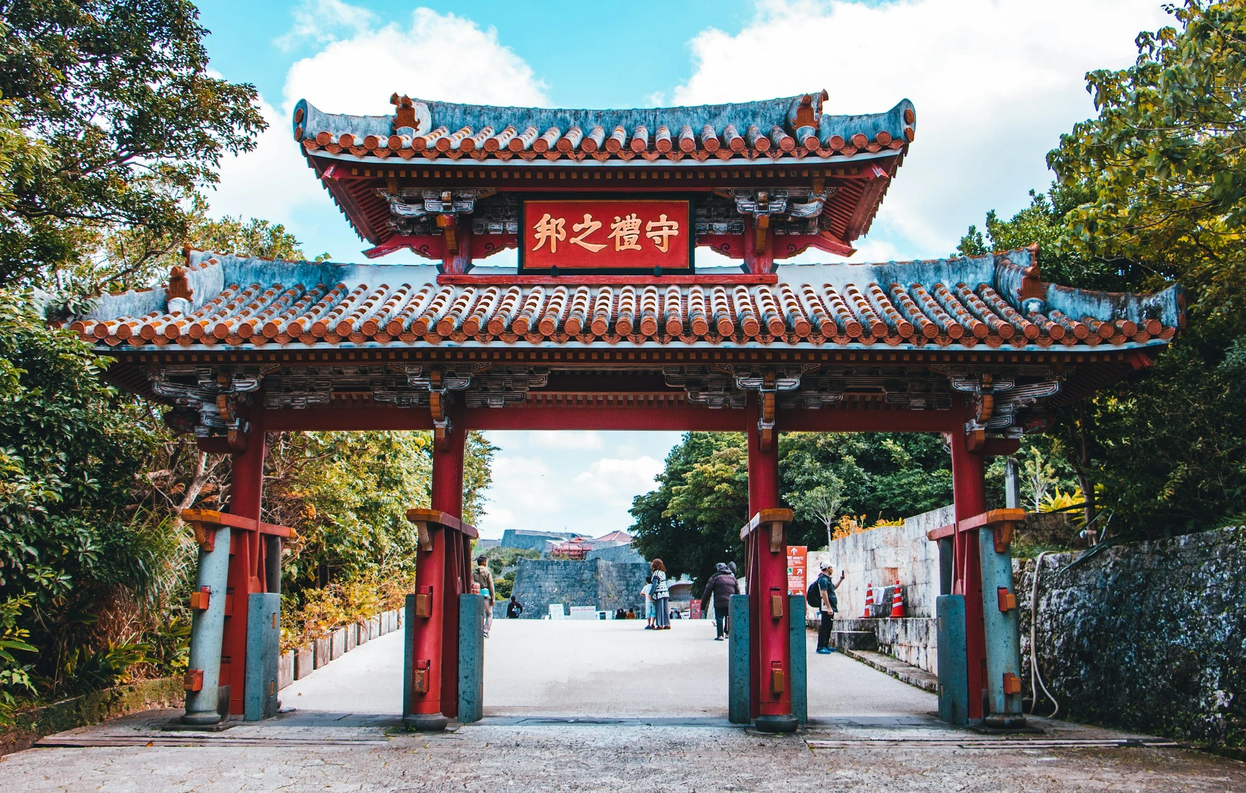 Traditional Asian-style gate with red columns, ornate roof, and a red sign with Chinese characters, surrounded by greenery and trees, with people walking underneath.