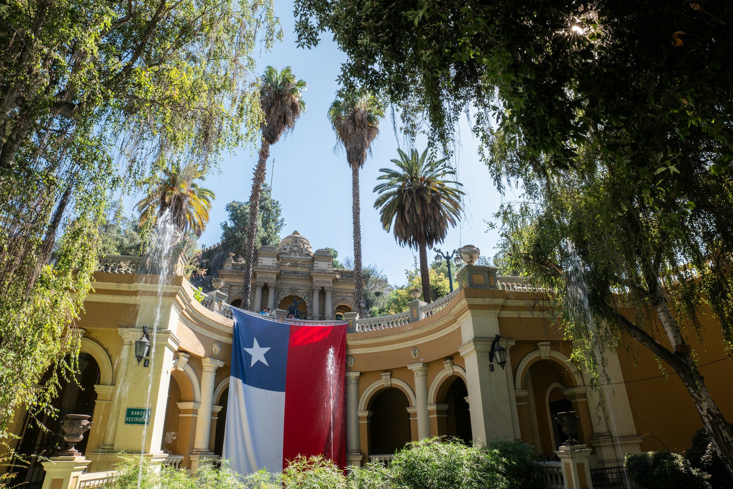 Tall palm trees and lush green trees surround a historic, ornate building with a Spanish flag hanging in front. Bright sunlight filters through the trees, creating shadows.