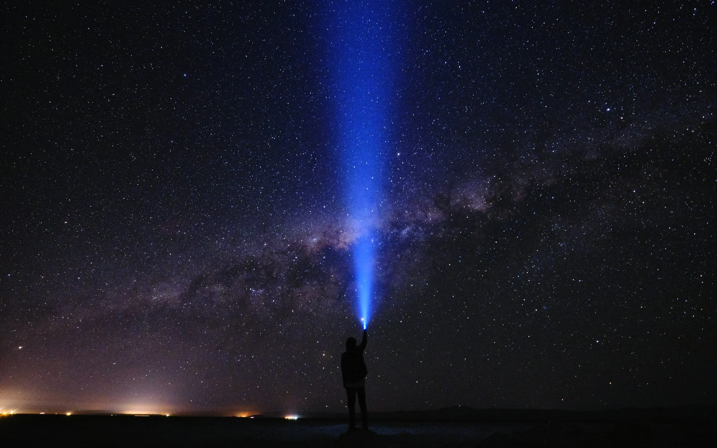 Silhouette of a person holding a blue flashlight directed at the night sky filled with stars and the Milky Way galaxy.