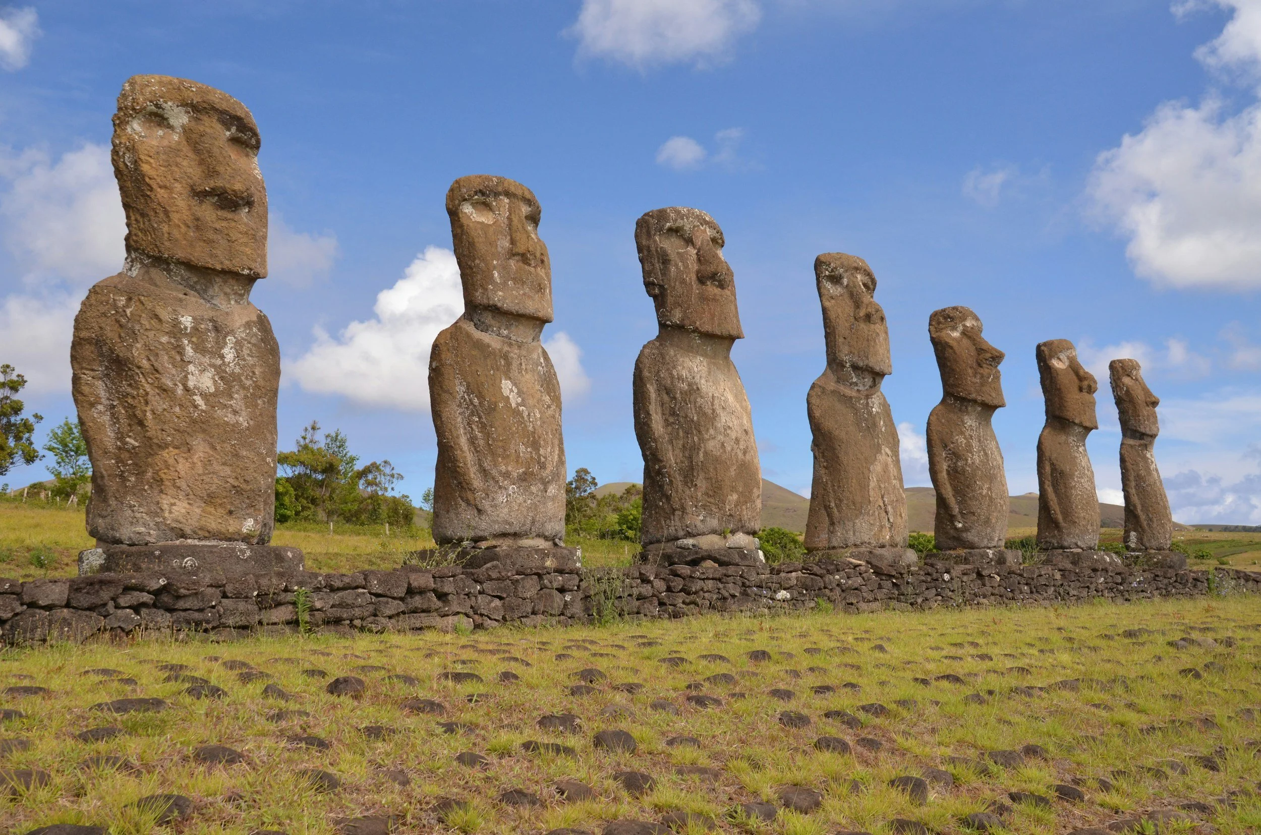 Line of Moai statues on a grassy landscape under a blue sky with scattered clouds.