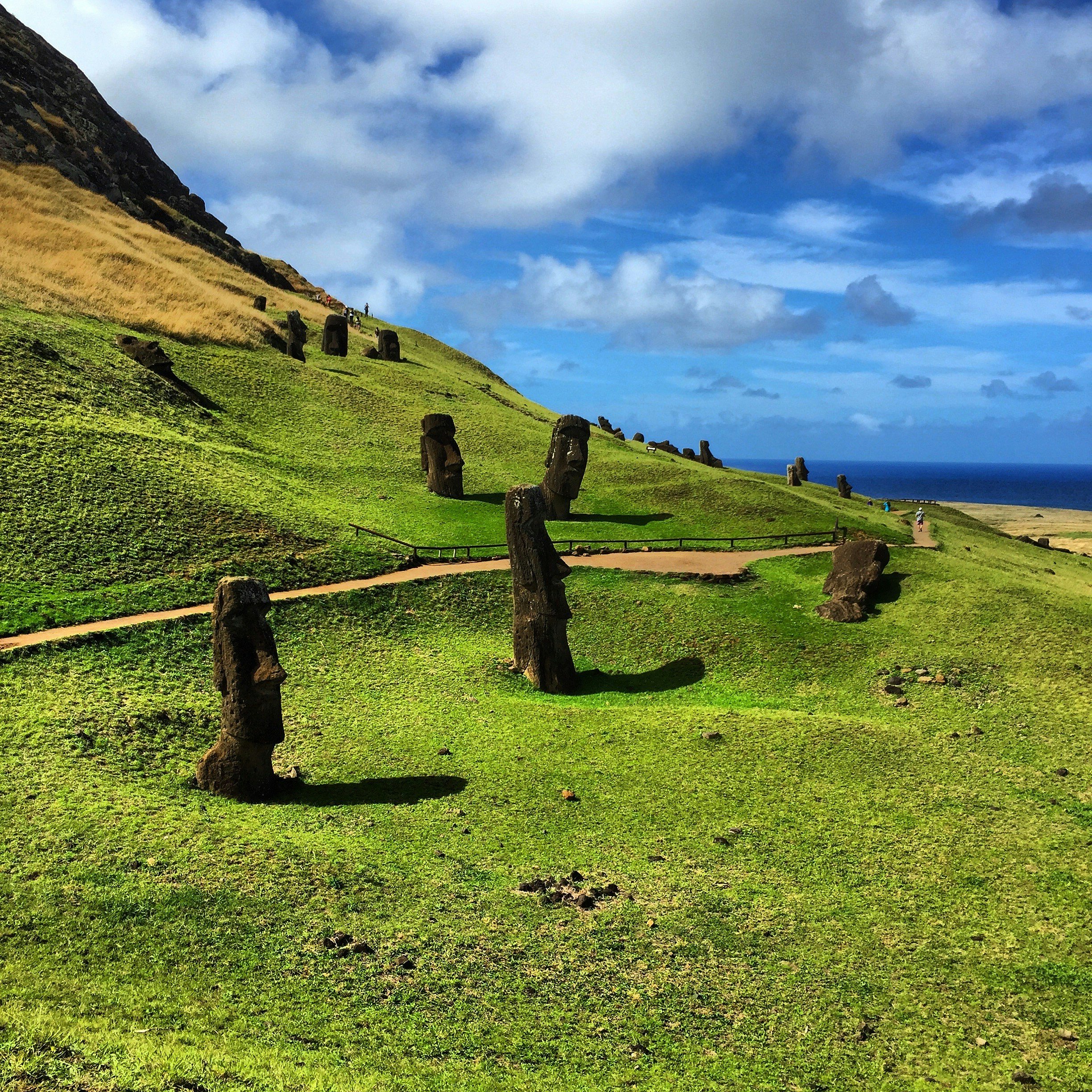 A grassy hillside with stone statues along a pathway, under a partly cloudy sky.