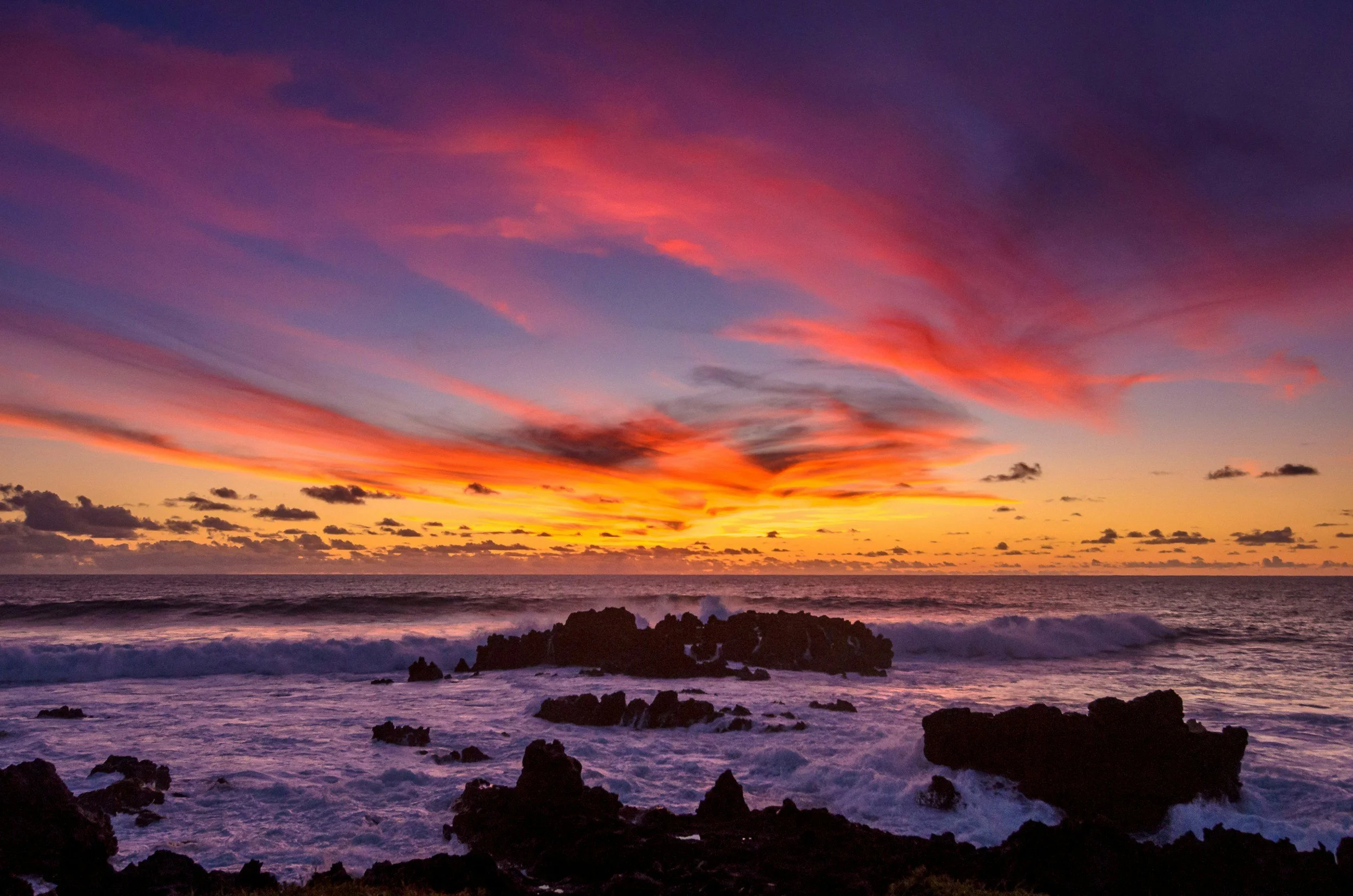 A colorful sunset over the ocean with pink, purple, and orange clouds, and dark rocks along the shoreline.
