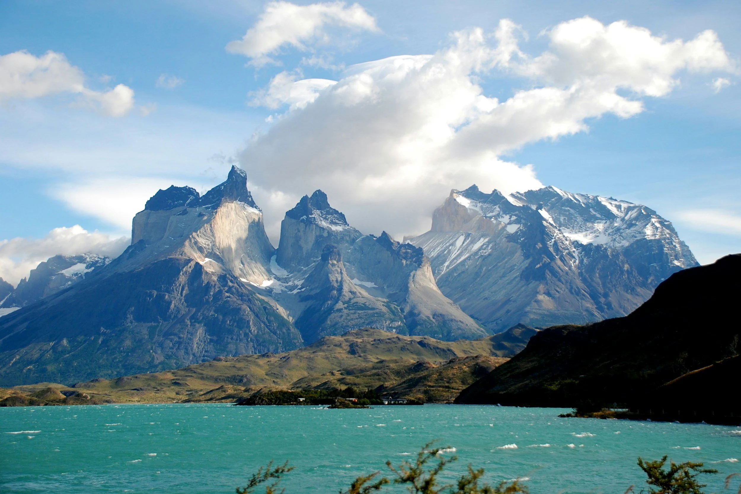 A landscape of a mountain range with snow-capped peaks, a large body of turquoise water in the foreground, and a partly cloudy sky.