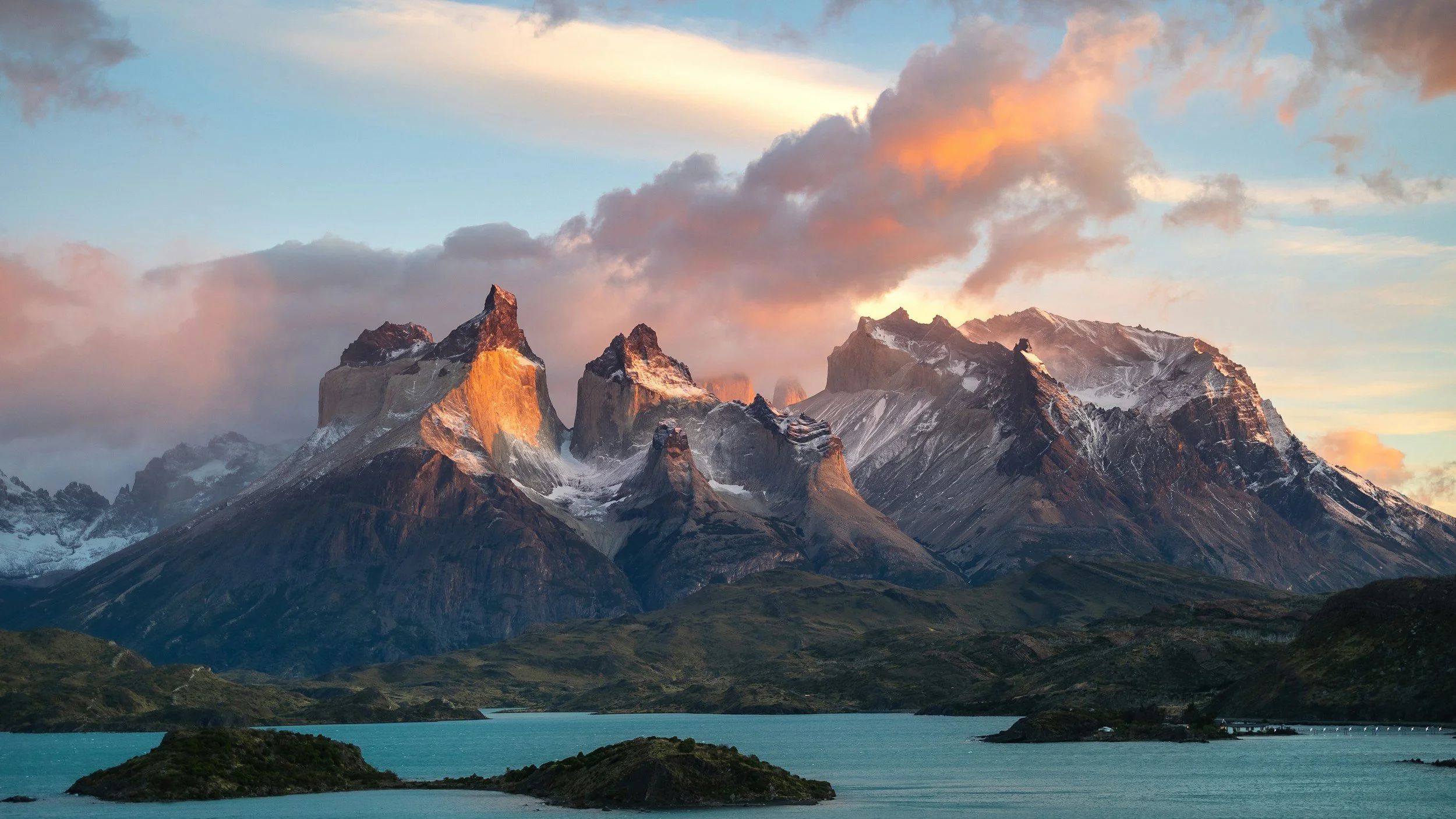 Snow-capped mountain peaks at sunset, with clouds in the sky and a turquoise lake in the foreground.