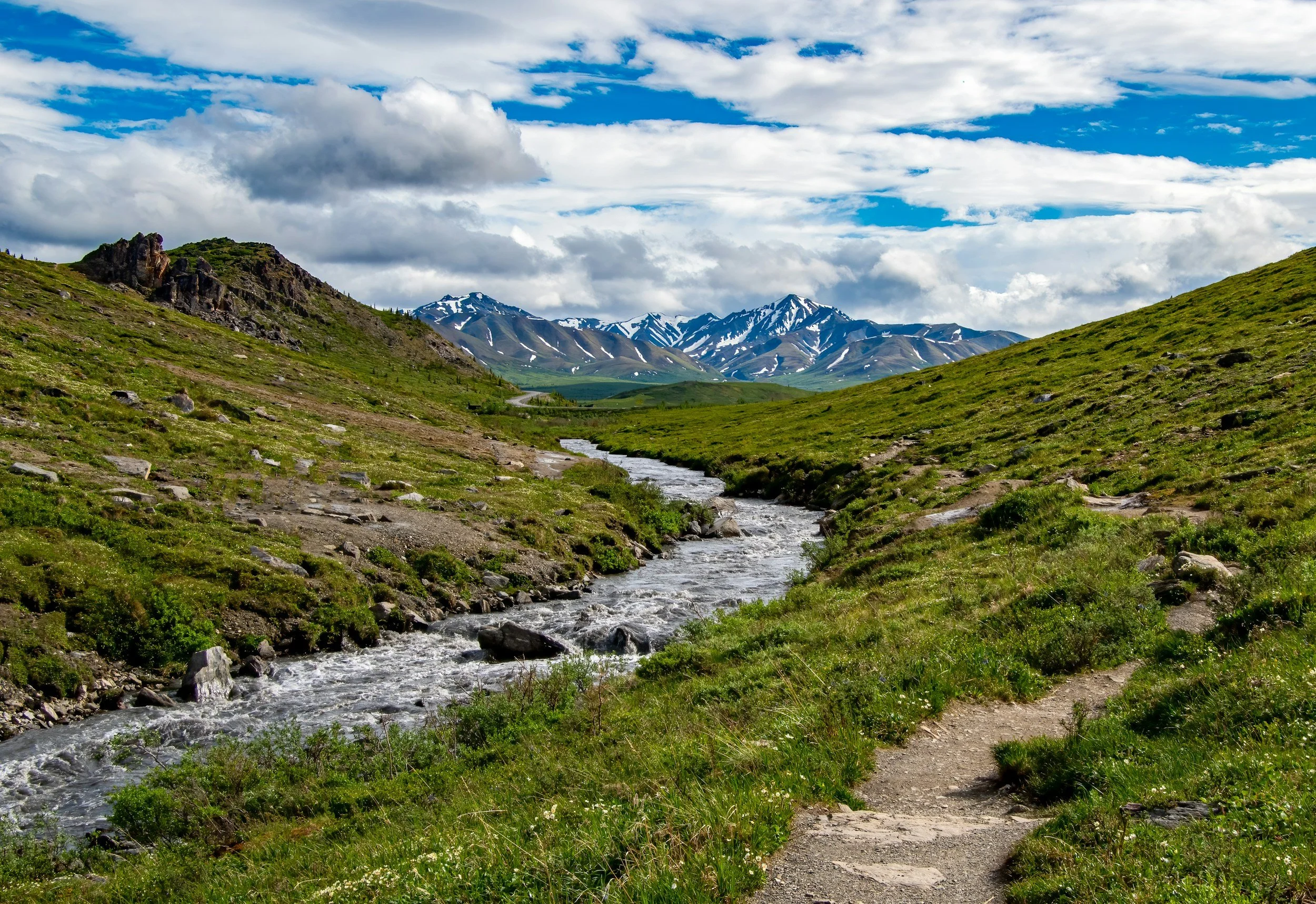 A scenic mountain landscape with a flowing creek in the foreground, green grassy hills, and snow-capped mountains under a partly cloudy sky.