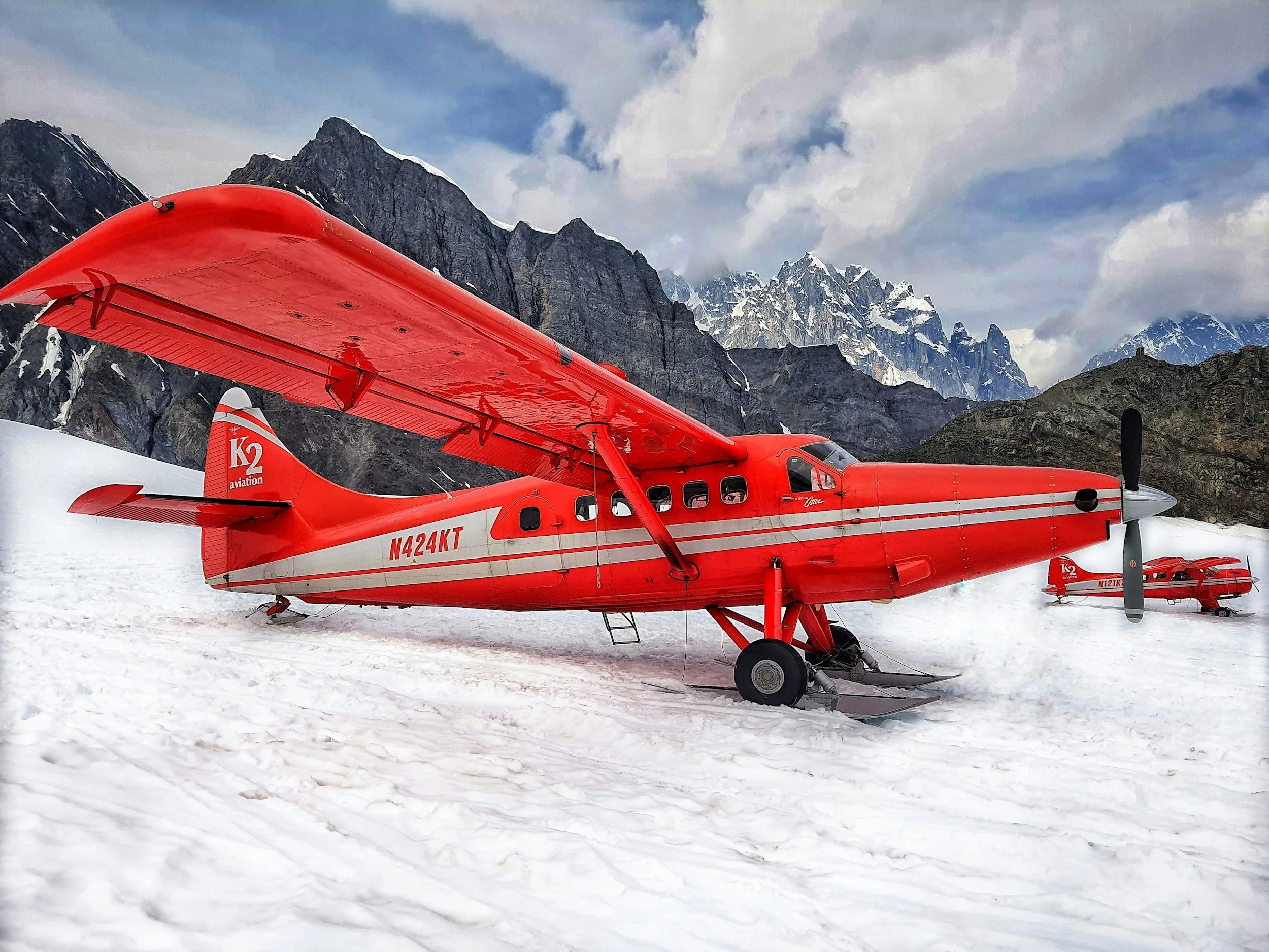 Red seaplane with skis on snow, mountains in background, partly cloudy sky.