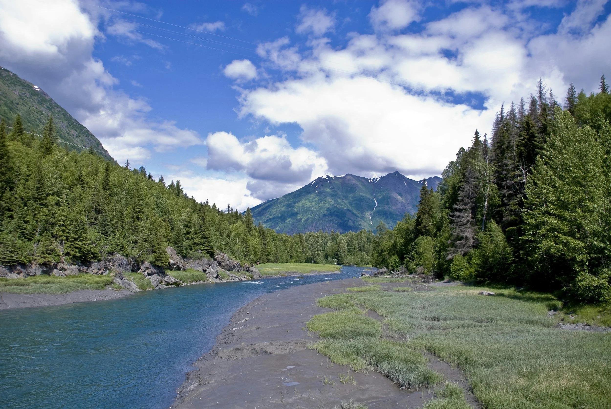 A scenic view of a river flowing through a forested valley with mountains in the background and a partly cloudy sky overhead.