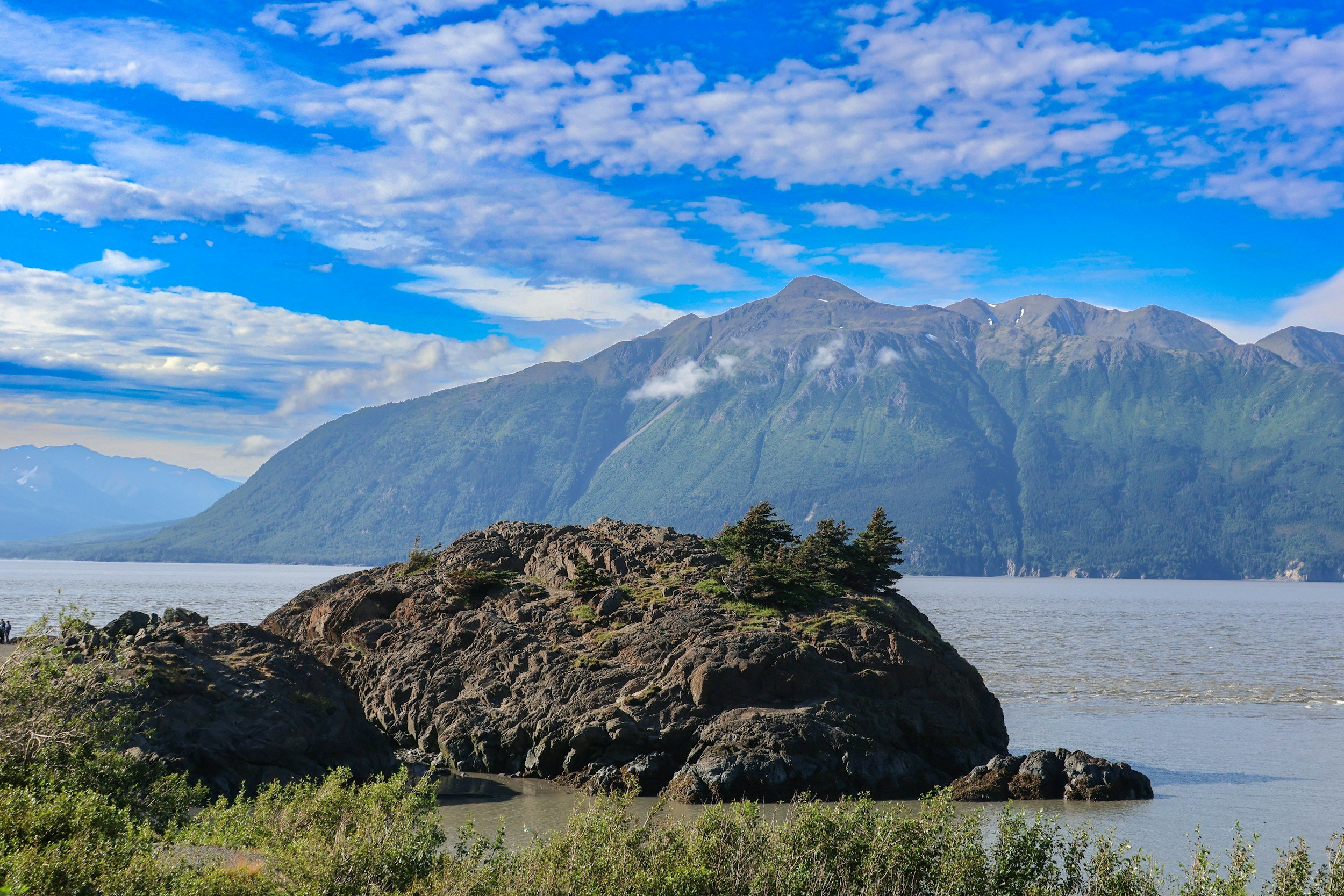 A scenic view of a mountain range with snow patches, partly covered with clouds, beside a body of water with rocky shoreline and green vegetation in the foreground.