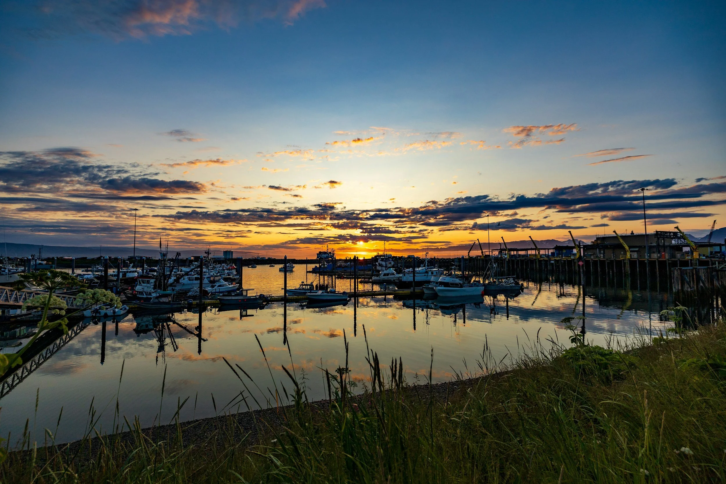 Sunset over a marina with boats docked in calm water, reflecting the colorful sky with orange and blue hues, and clouds scattered across the horizon.
