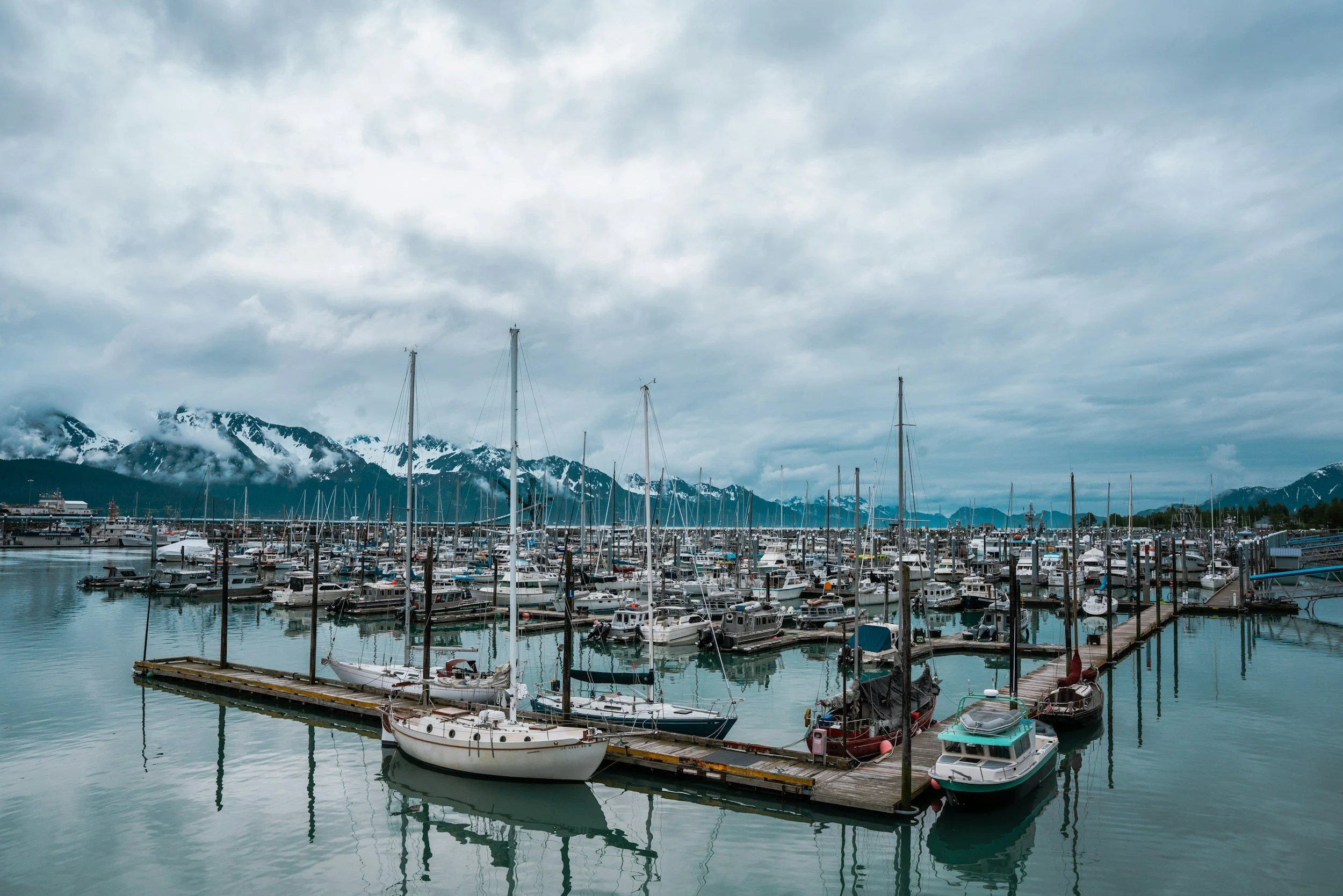 A marina with numerous boats docked, snow-capped mountains in the background, and a mostly cloudy sky overhead.