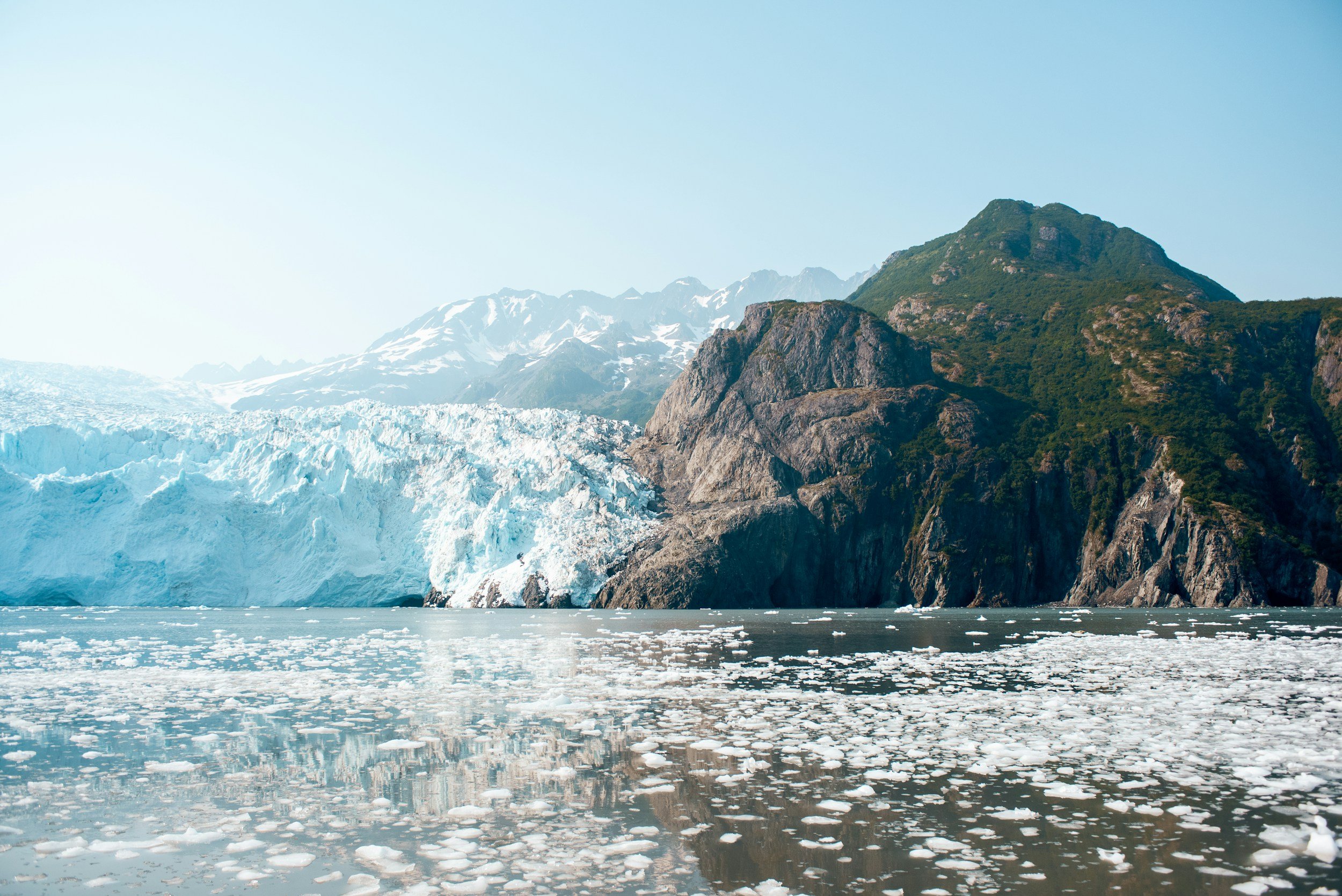 Glacier and snow-covered mountains along a rocky coastline with floating ice in the water.