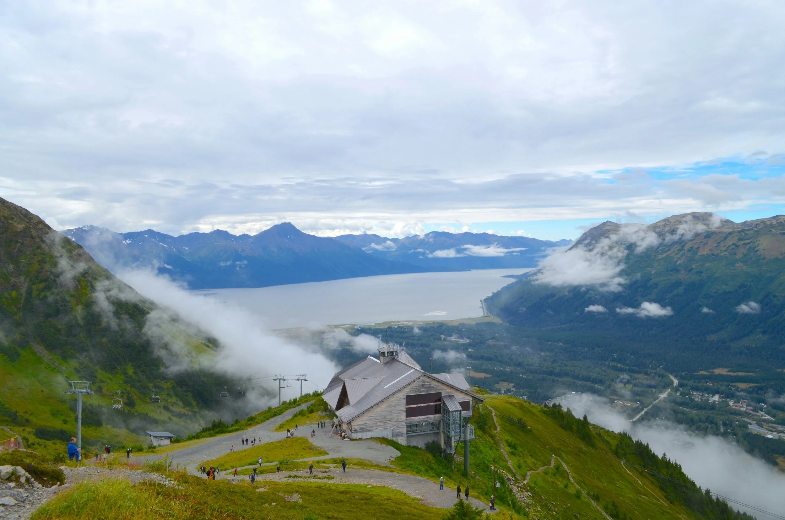 Mountain landscape with a lake in the valley, clouds partly covering the peaks, and a building in the foreground with people walking around.