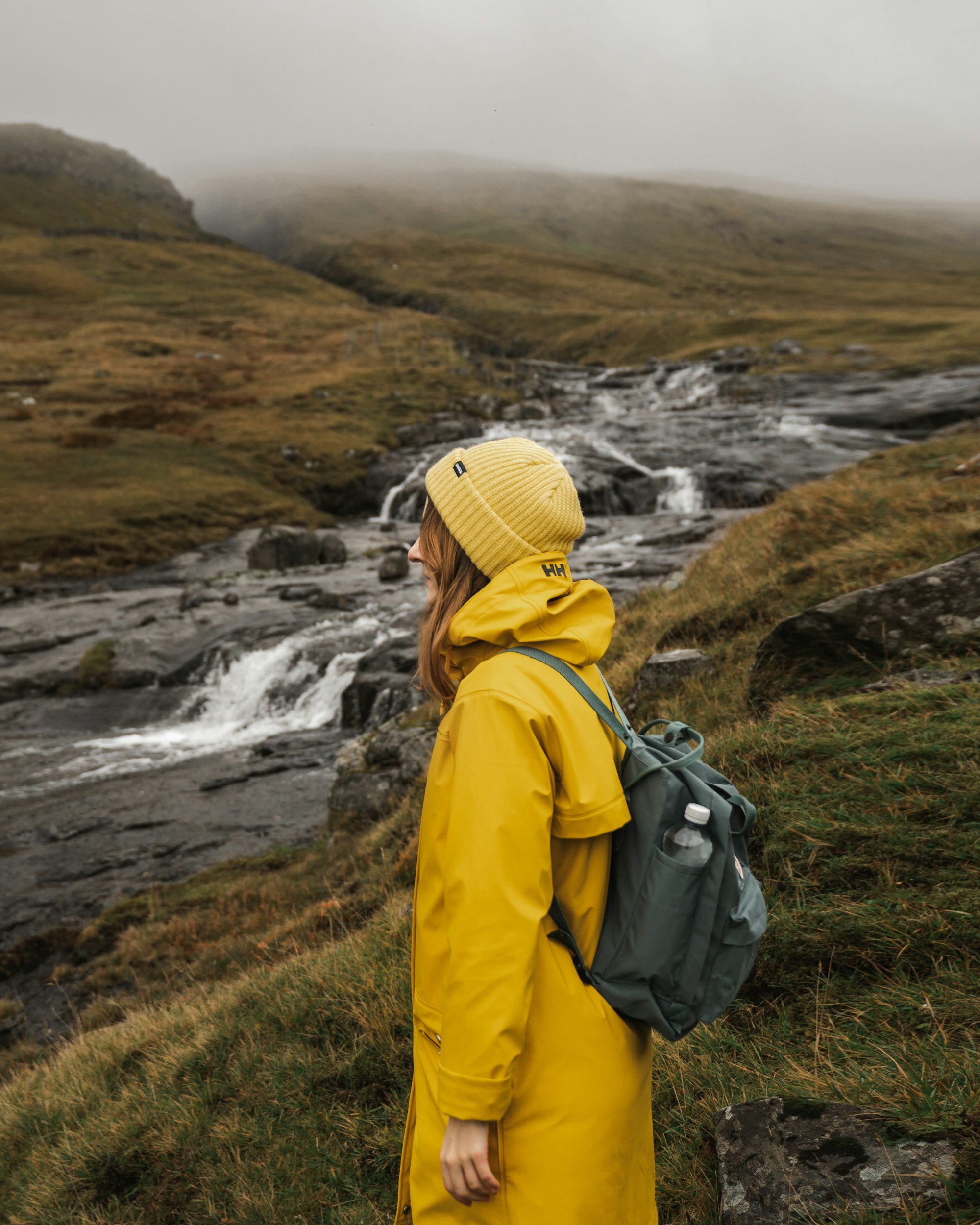 A person wearing a yellow beanie and a yellow raincoat carrying a gray backpack with a water bottle, standing outside near a flowing stream in a mountainous area with low clouds and green hillside.