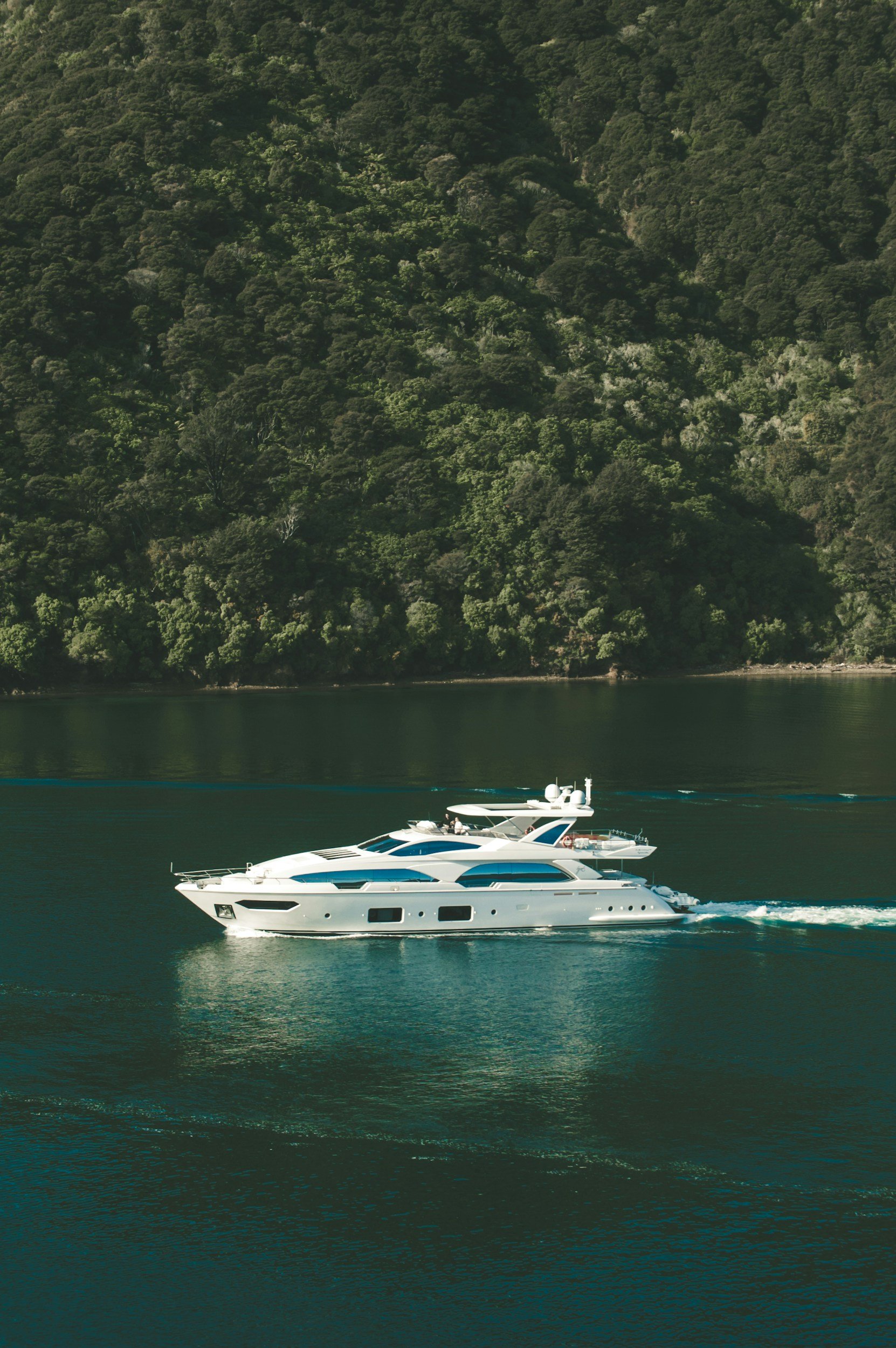 A white yacht sailing on calm water near a green forested hillside.