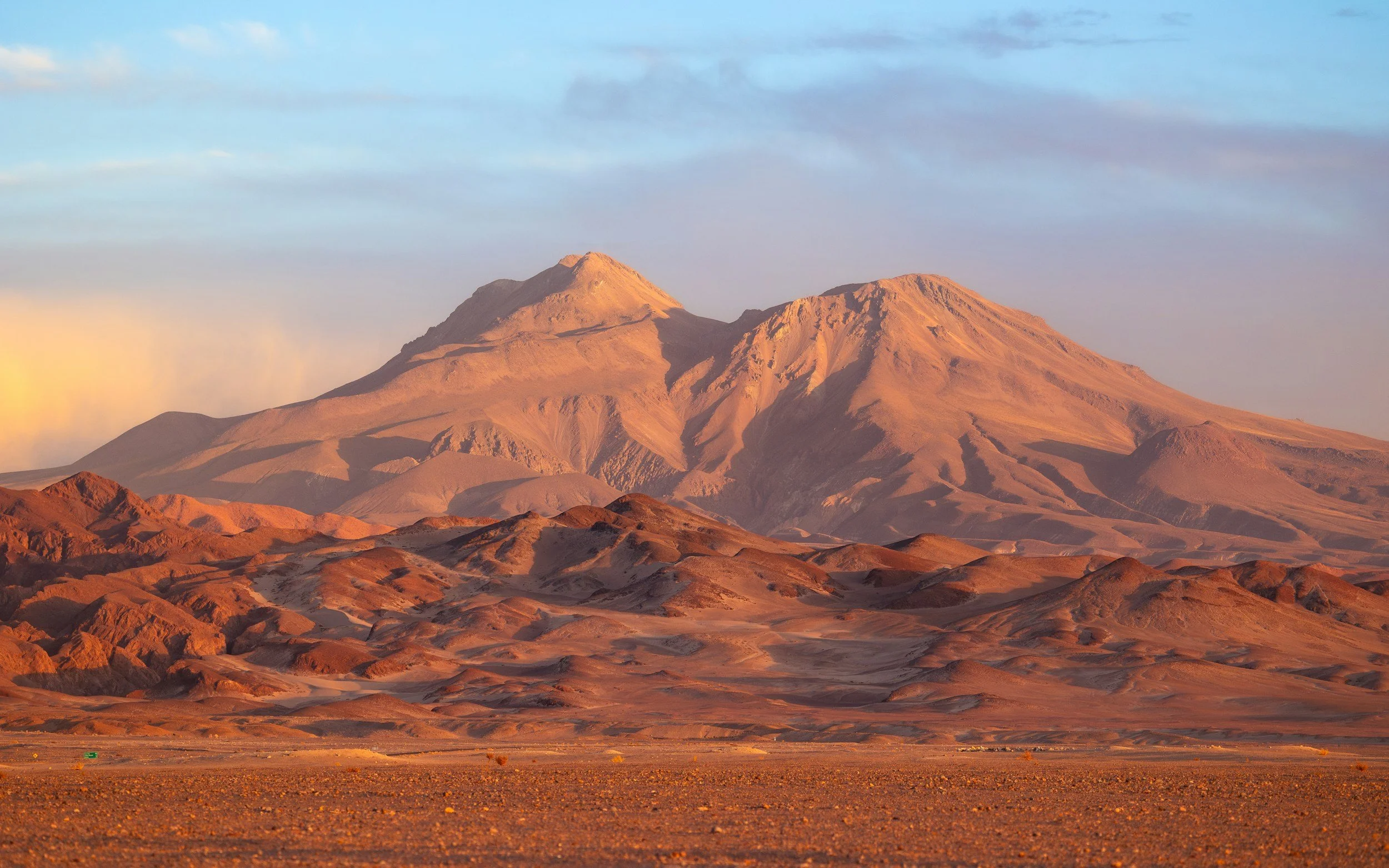 Sunset over rugged desert mountains with reddish-brown tones and expansive blue sky.