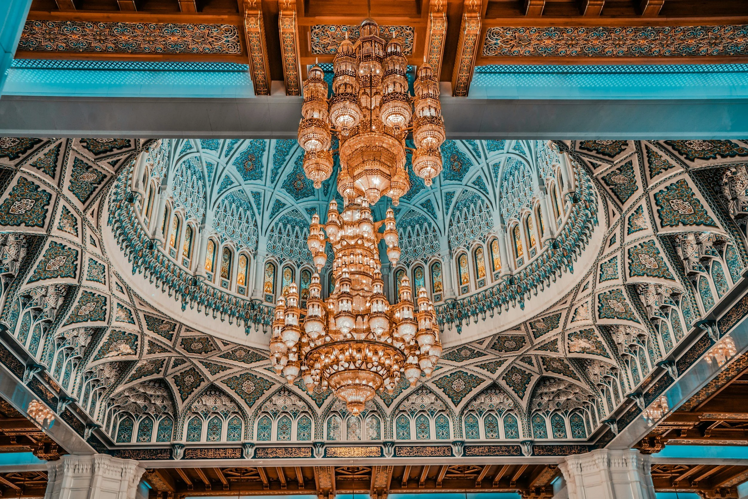 An ornate interior featuring a large golden chandelier hanging from intricately decorated ceilings with Islamic geometric patterns and arches in a mosque or palace.