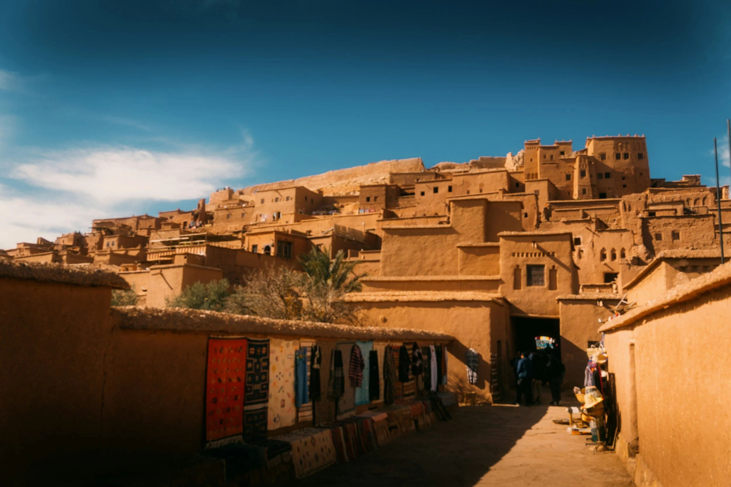A hillside city with traditional earthen buildings under a blue sky, with a market stall with colorful textiles and people walking in the street.