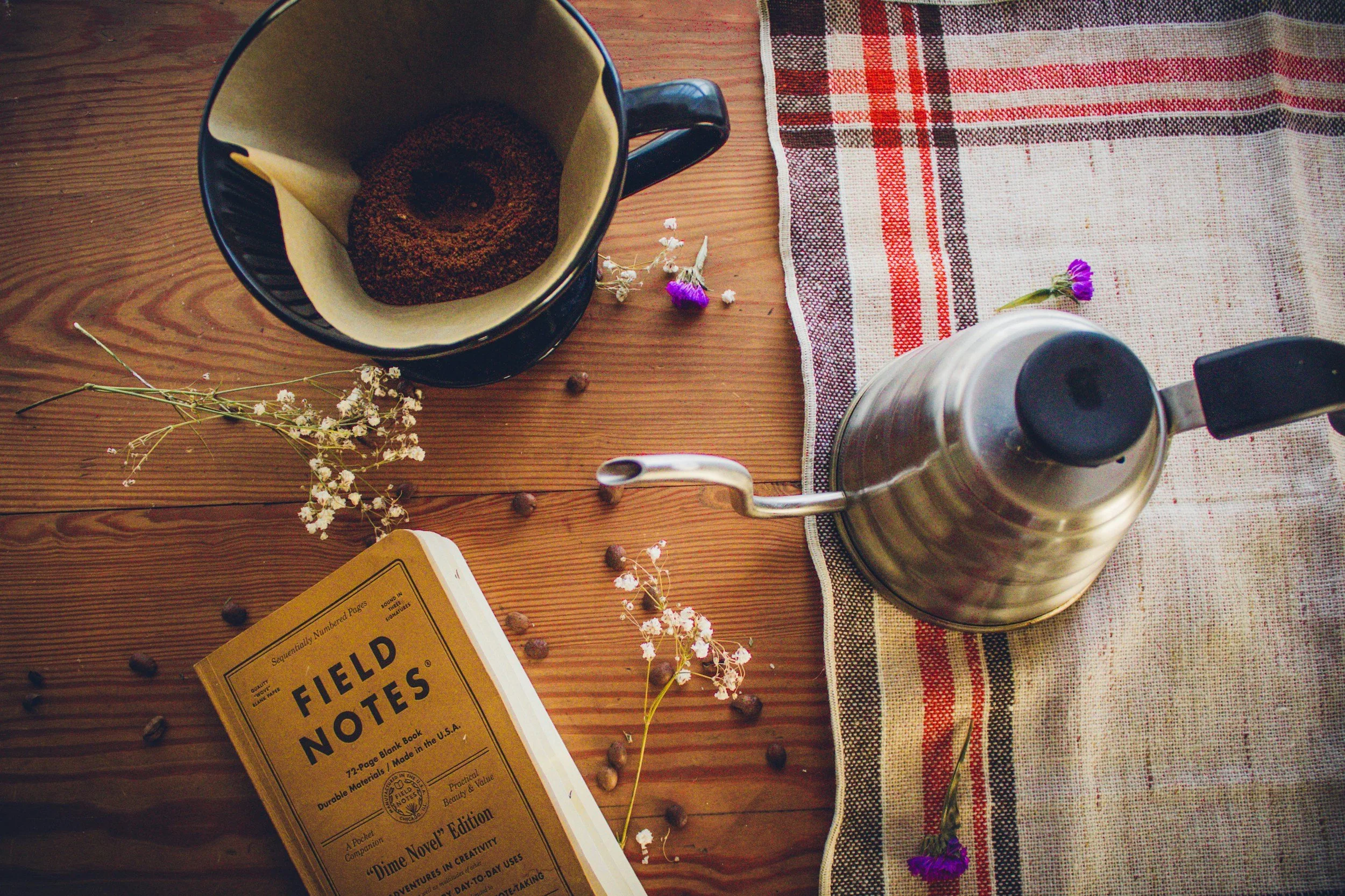 Top down view of a wooden table with a black coffee mug containing coffee filters, a metal kettle, a cloth with red, black, and white stripes, scattered small purple and white flowers, a yellow book, and some scattered coffee beans.