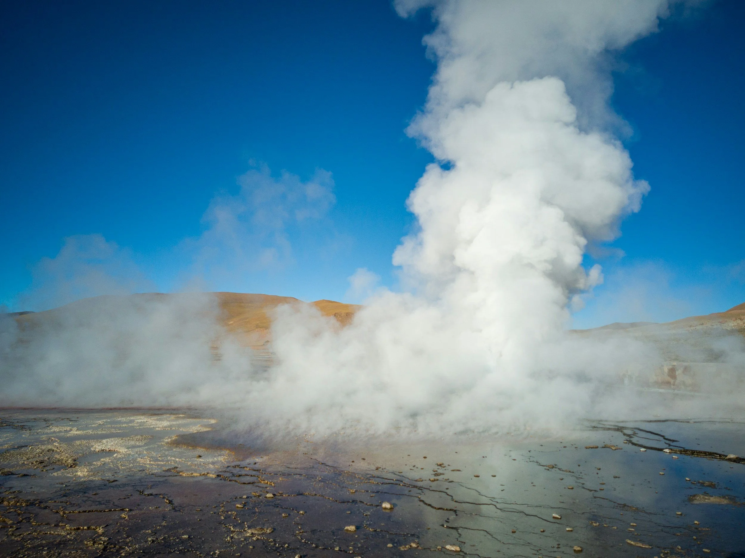 A tall column of steam rising from the El Tatio geyser field against a bright blue sky.