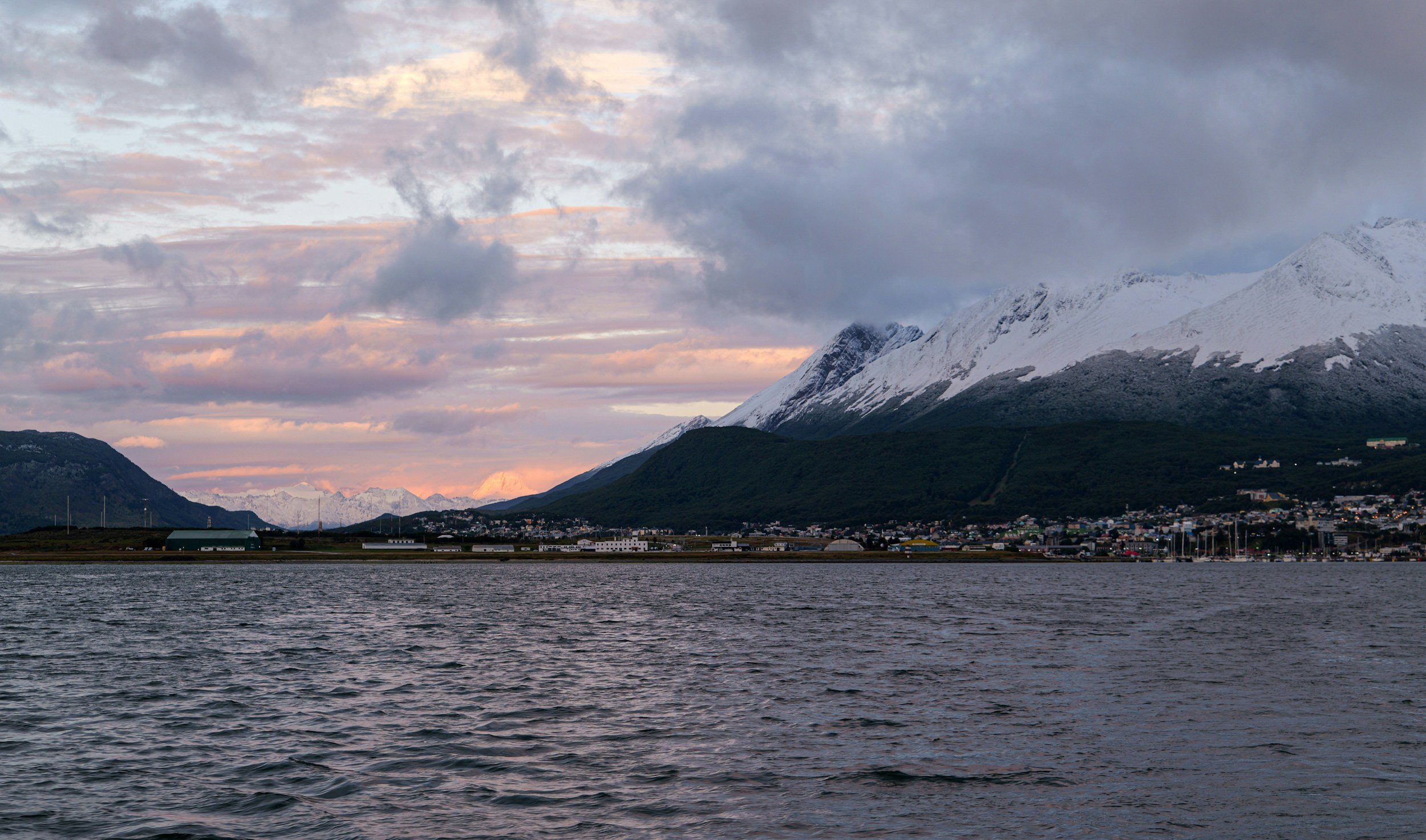 A snowcapped mountain glowing pink at dusk above the Beagle Channel and the lights of Ushuaia.