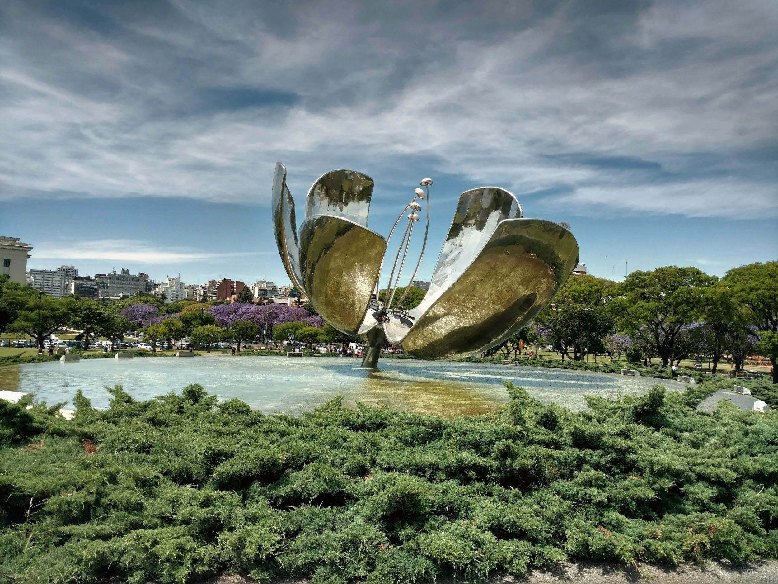 The giant steel flower sculpture Floralis Genérica opening above a reflecting pool, with purple jacaranda trees in bloom in Buenos Aires.