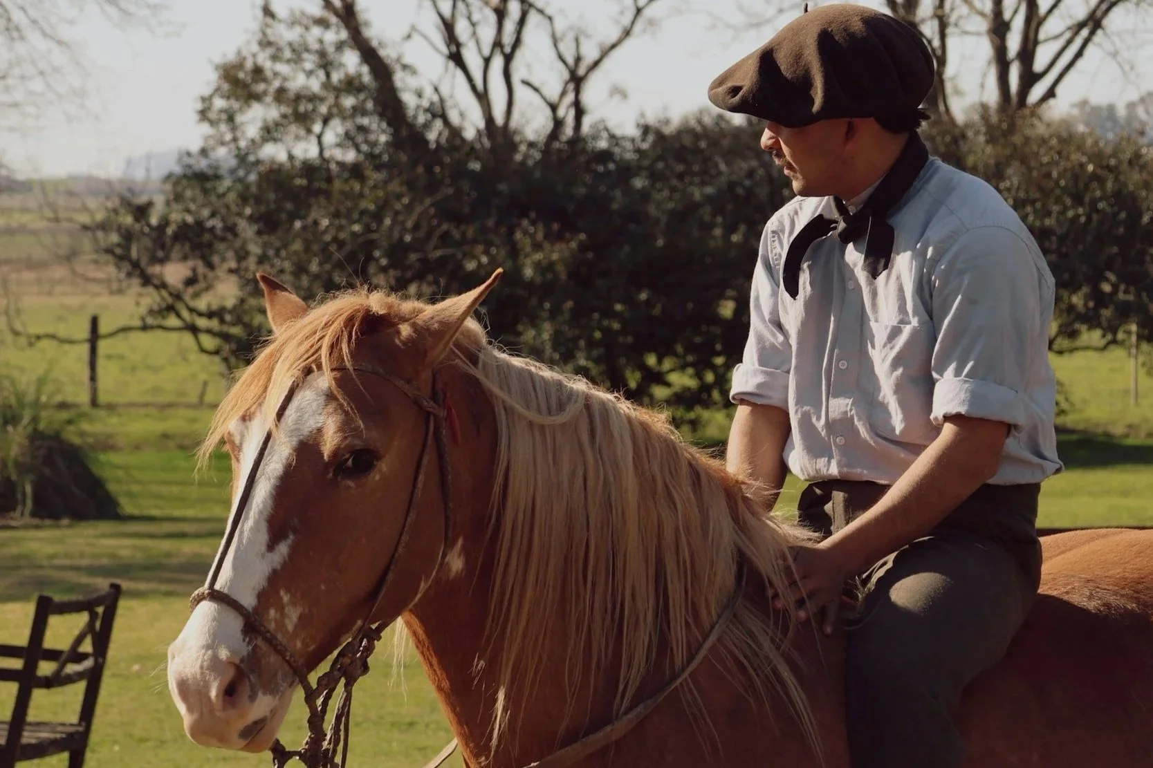 A gaucho in a flat cap seated on a chestnut horse with a pale mane on a grassy estancia.