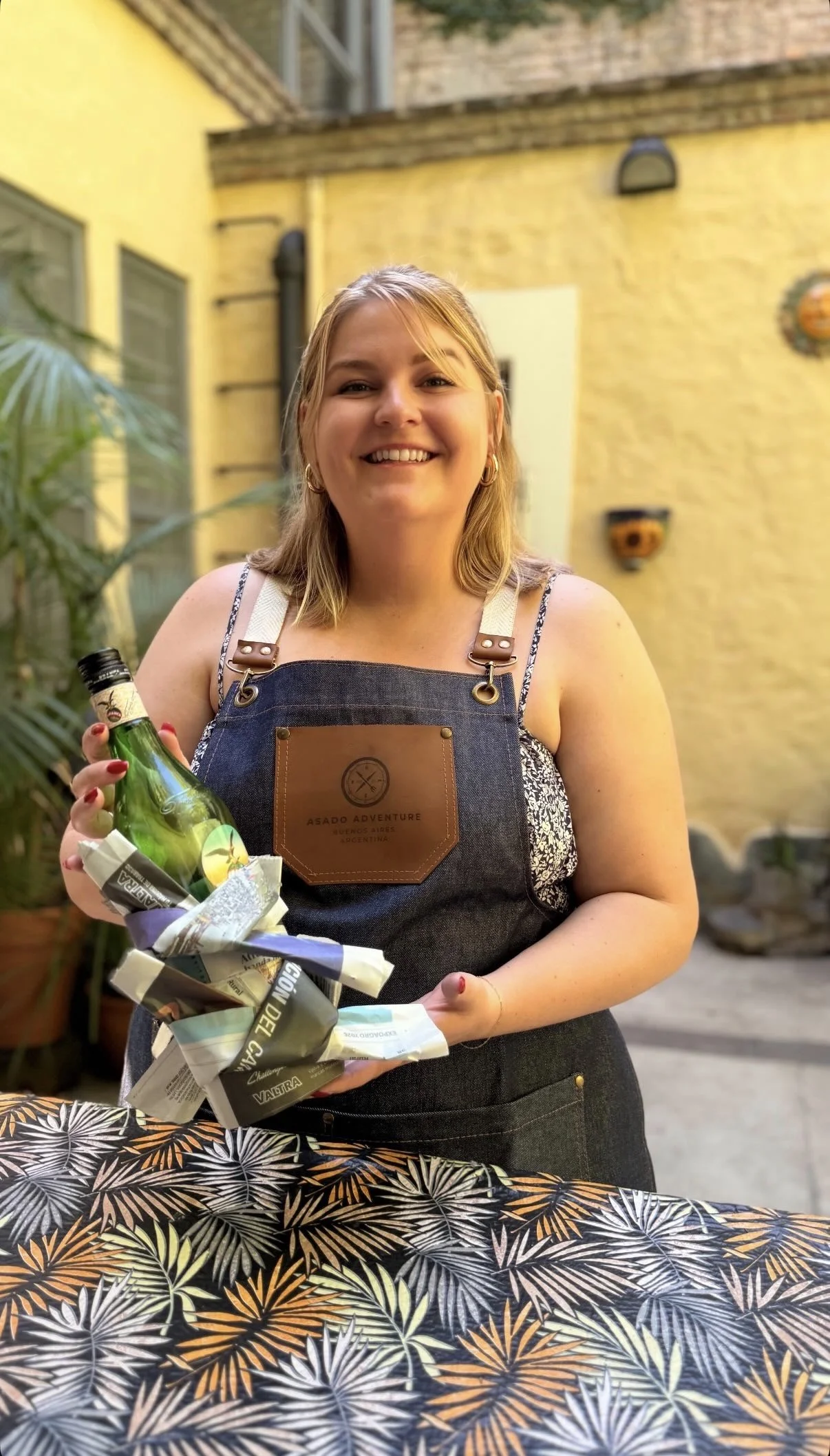 Smiling woman wearing a denim apron holding a tray with a bottle and wrapped food items outside on a sunny patio.