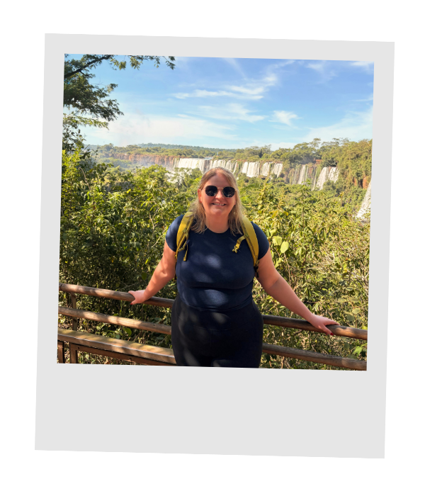 Piper smiling at a railing above Iguazú Falls with rainforest and waterfalls behind her.