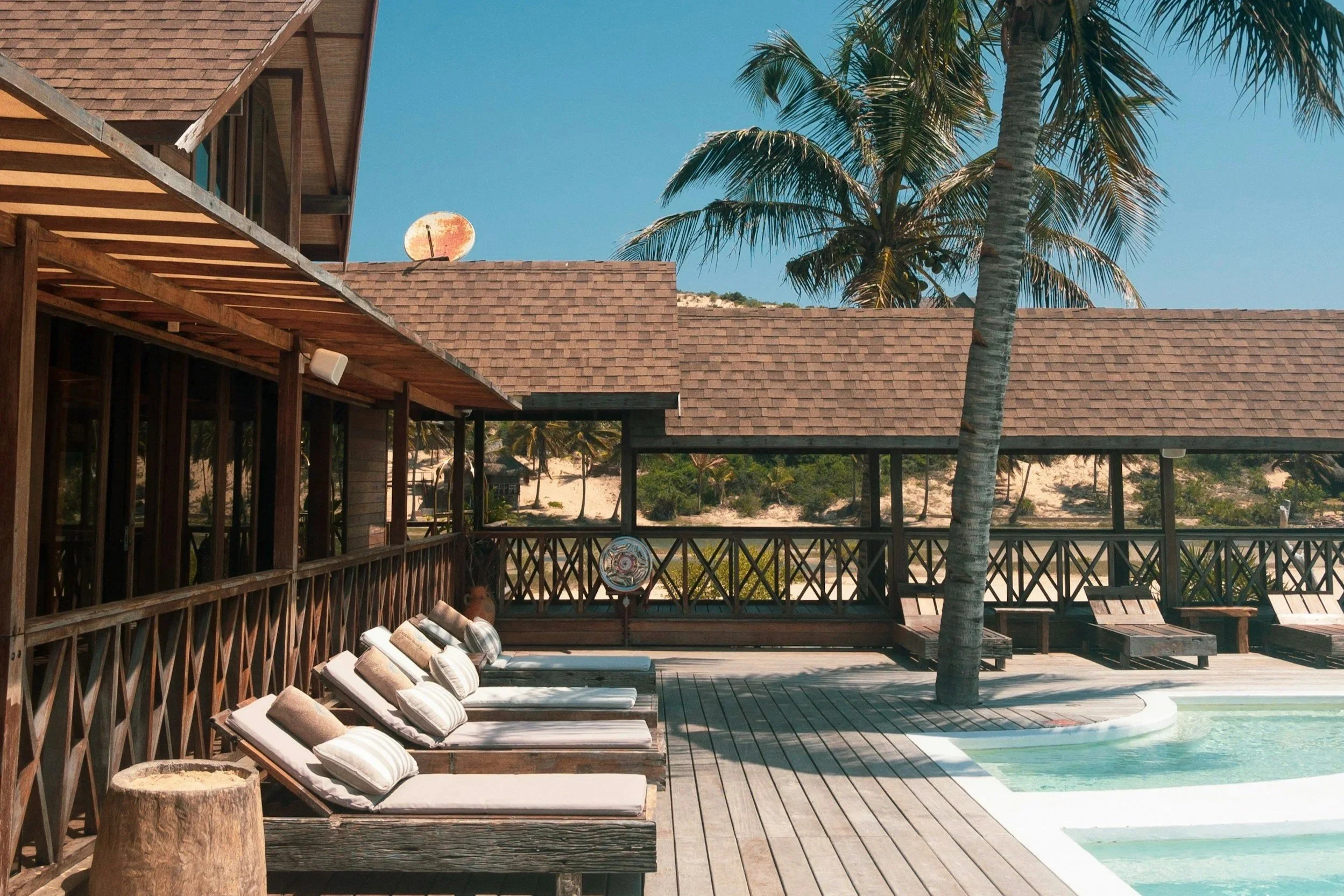 A wooden deck outdoor area with lounge chairs and cushions next to a swimming pool, with a palm tree and house roof in the background under a blue sky.