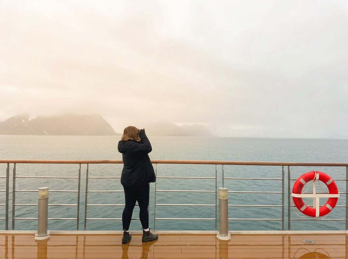 A person standing on a boat deck taking a photo of the distant mountains and the ocean with a camera, wearing a black jacket and pants, with a life preserver on the railing nearby.