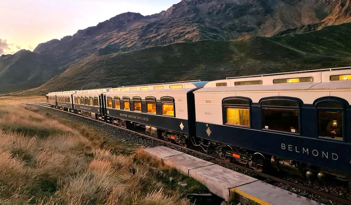 The Belmond Andean Explorer luxury train crossing golden high-altitude grassland at sunset, its cabin windows glowing.