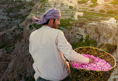 Man wearing a headscarf holding a basket filled with pink flowers, overlooking a hilly landscape with buildings.