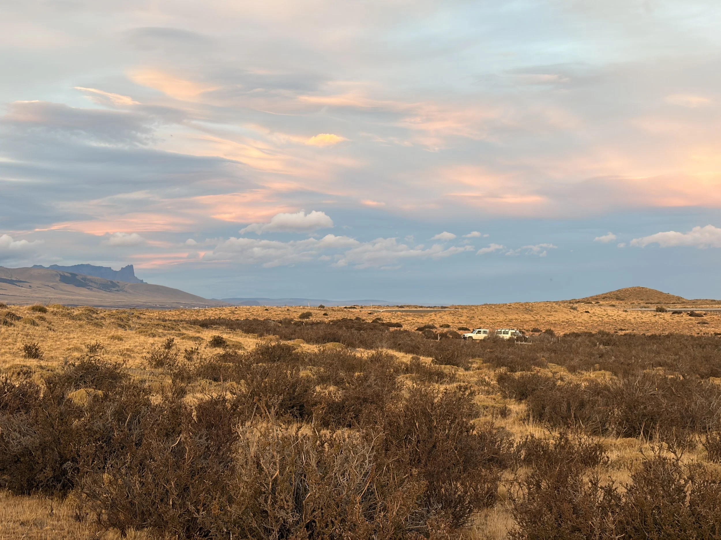 Golden Patagonian grasslands stretching toward distant flat-topped mountains, with two white expedition vehicles parked along a dirt road.