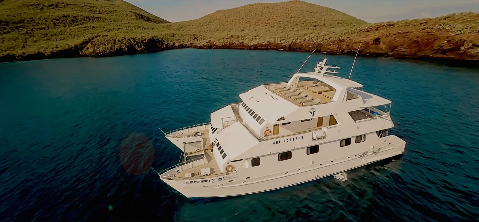 A white private yacht anchored in a deep blue cove beneath green and rust-colored volcanic hills in the Galápagos