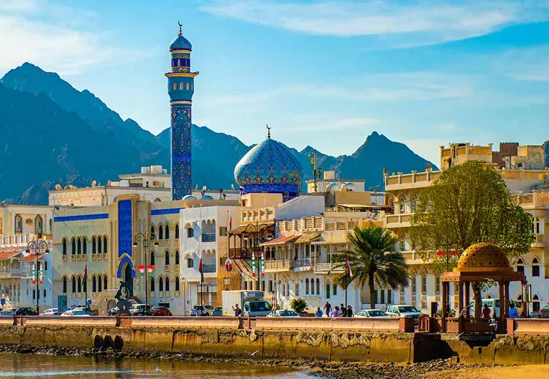 A picturesque waterfront view of a city with a mosque featuring a blue dome and a tall minaret, surrounded by buildings, palm trees, and mountains in the background.
