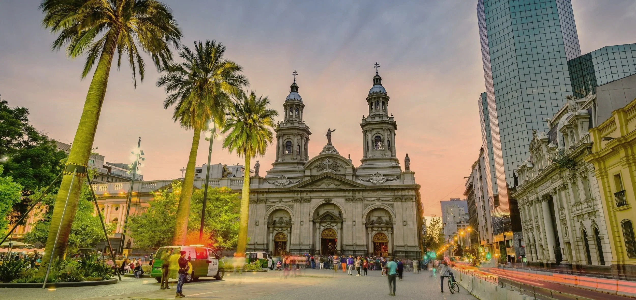 The twin towers of Santiago's Metropolitan Cathedral glowing at dusk above palm trees and evening crowds in the Plaza de Armas.